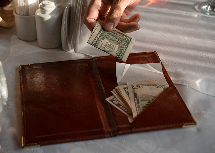 Hand placing cash tip into bill folder on table, illustrating tipping a waitress on a 300 plus tab debate. Hand placing cash tip into bill folder on table, illustrating tipping a waitress on a 300 plus tab debate.