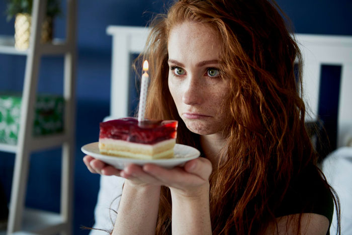 An anxious birthday girl holding a small cake with a lit candle, looking overwhelmed during surprise bash with 50 guests.
