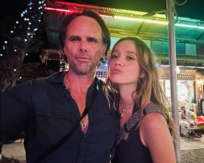 Walton Goggins and Aimee Lou Wood posing together outdoors at night with lights and a building in the background