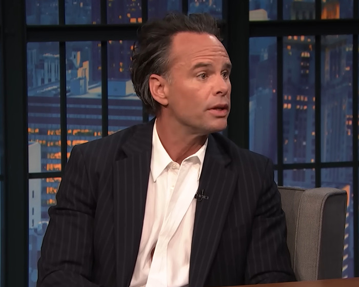 Walton Goggins in a dark pinstripe suit during a late night talk show interview, reacting to feud questions.