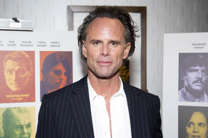 Walton Goggins at an event, dressed in a dark pinstripe suit and white shirt, with yearbook photos displayed behind him.