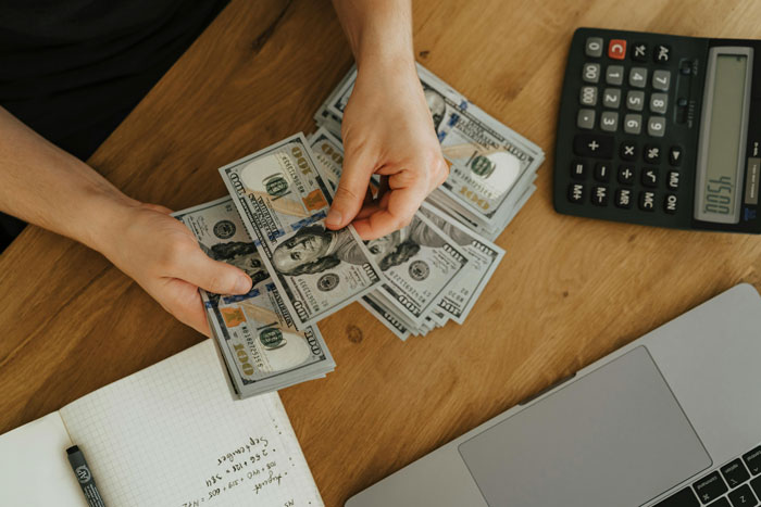 Hands counting US dollar bills next to a calculator and laptop, illustrating client fails to pay and freelancer tricks.