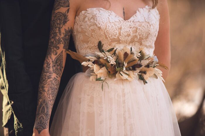 Bride in a lace wedding dress with tattooed arm holding a dried floral bouquet, symbolizing soul-crushing parental words.
