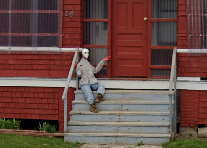 Alien-like figure sitting on porch steps of a red house, one of the weird things on Google Earth that will make you question reality.