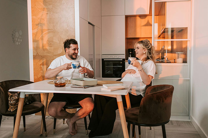 Man and wife laughing and drinking tea at a modern kitchen table, capturing a light moment in their relationship.