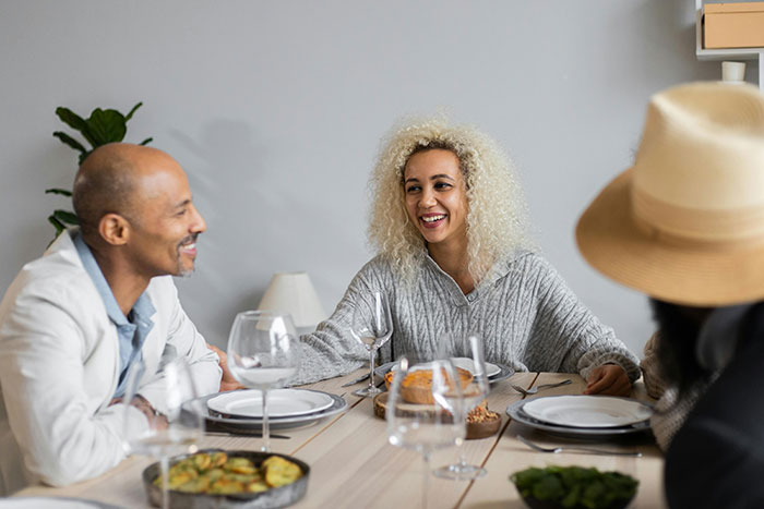 Man smiling at woman with curly hair at dining table with wife&rsquo;s best friend, capturing a tense social moment.