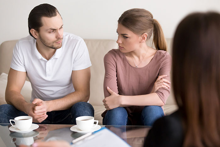 Man and wife sitting on couch looking upset during a tense conversation with a third woman present.