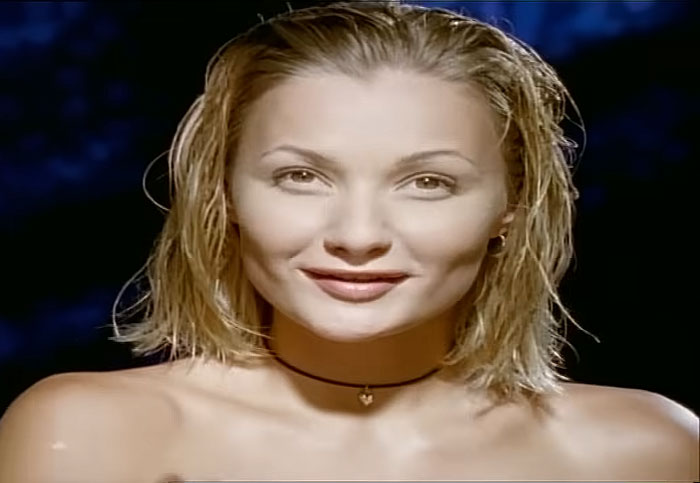 90s megastar Whigfield smiling with wet hair and natural makeup in a close-up portrait against a dark background.