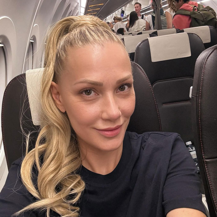 90s megastar Whigfield with flawless skin and a high ponytail, sitting inside an airplane cabin, smiling at the camera