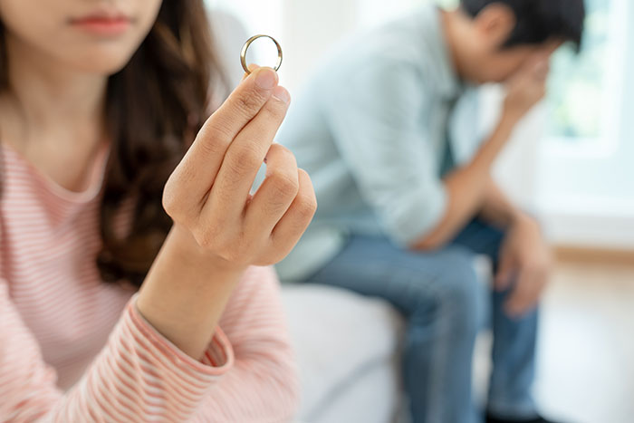 Wife holding wedding ring looking upset, husband sitting in background with head in hands, emotional conflict visible.