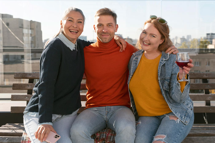 Three friends sitting close together on a bench outdoors, a woman holding a wine glass, smiling and enjoying a casual gathering.