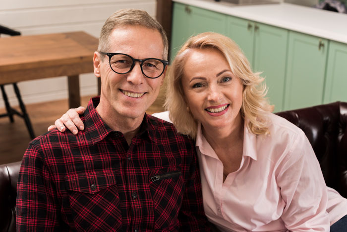 Middle-aged couple smiling together on a couch, depicting a wife&rsquo;s catch-up with her ex and a husband home alone.