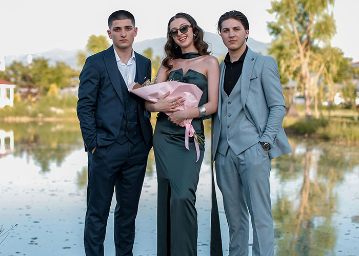 Three well-dressed young adults posing outdoors by water, representing a fake relationship and rumored cheating scenario.