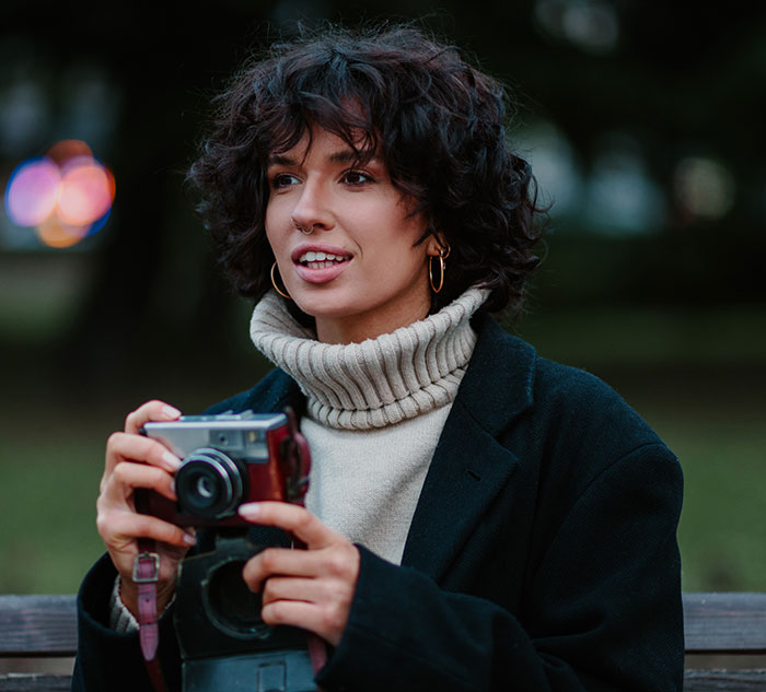 Young woman holding a camera outdoors, representing an entitled mom interrupting a photoshoot for free pics of her kids.