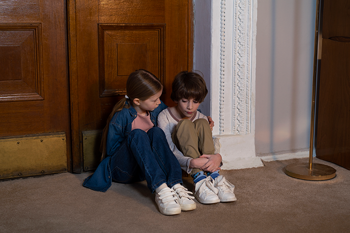 Two young siblings sitting on a floor inside an empty house, appearing sad and comforted by each other.