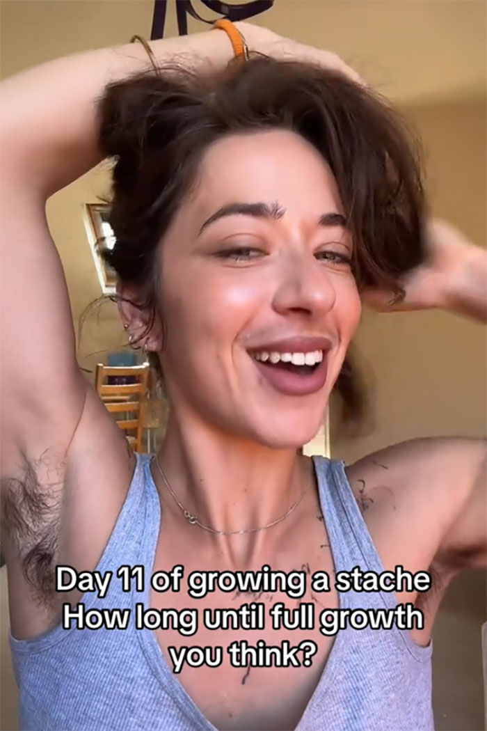 Woman challenges beauty stereotypes with natural facial and body hair, smiling confidently while raising arms indoors.