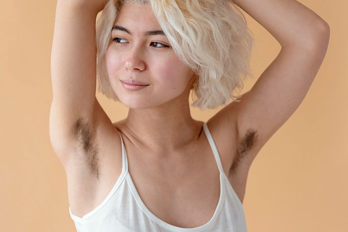 Woman challenges beauty stereotypes with natural armpit hair, confidently embracing her unique look against a neutral background.