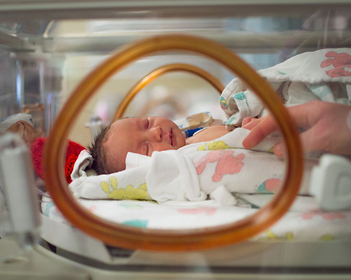 Newborn baby resting in a hospital incubator with a hand gently touching the blanket, highlighting family and care.