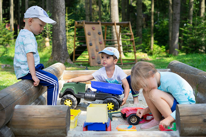 Three kids playing with toy cars in a sandbox outdoors, illustrating family drama and babysitting issues.