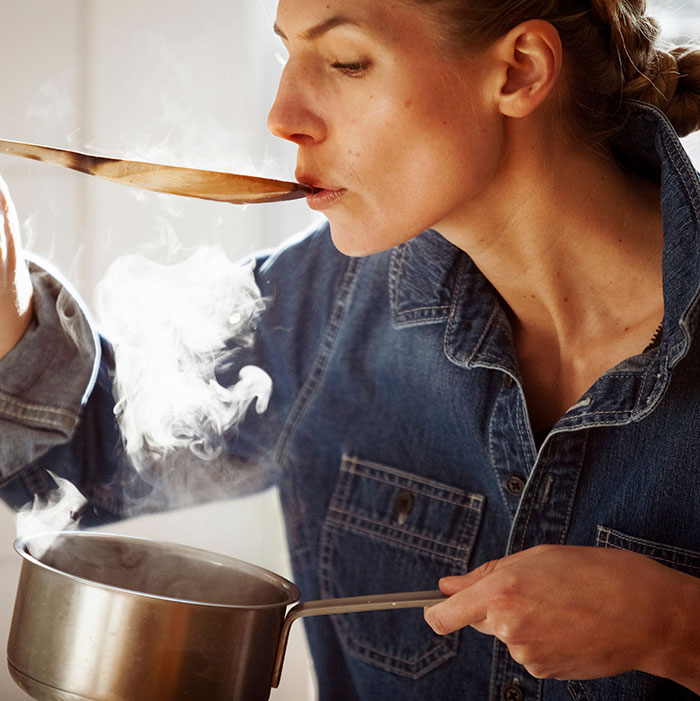 Woman in denim shirt tasting hot food from a wooden spoon over a steaming pot, illustrating Ozempic tongue side effects.