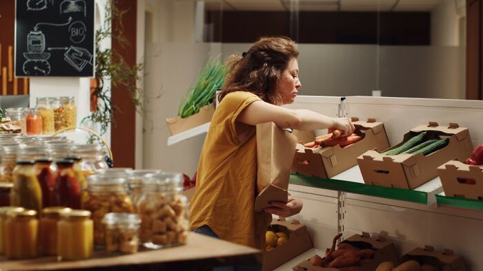 Woman selecting fresh vegetables from boxes in a store, with jars of preserved food in the foreground and a refrigerator nearby.