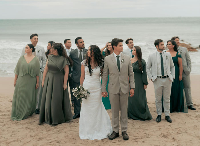 Bride and groom holding hands on beach with wedding party, highlighting fianc&eacute; at friend&rsquo;s wedding in formal attire.