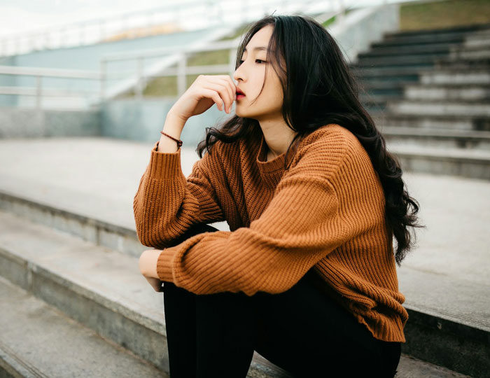 Young woman in a brown sweater sitting on outdoor steps, looking pensive about fianc&eacute; and friend&rsquo;s wedding situation.