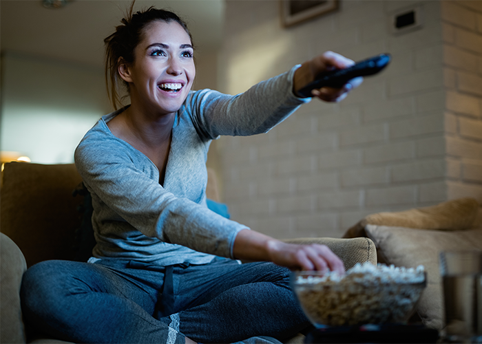 Woman smiling alone on couch in comfortable clothes, holding remote and reaching for popcorn, enjoying solitude at home.