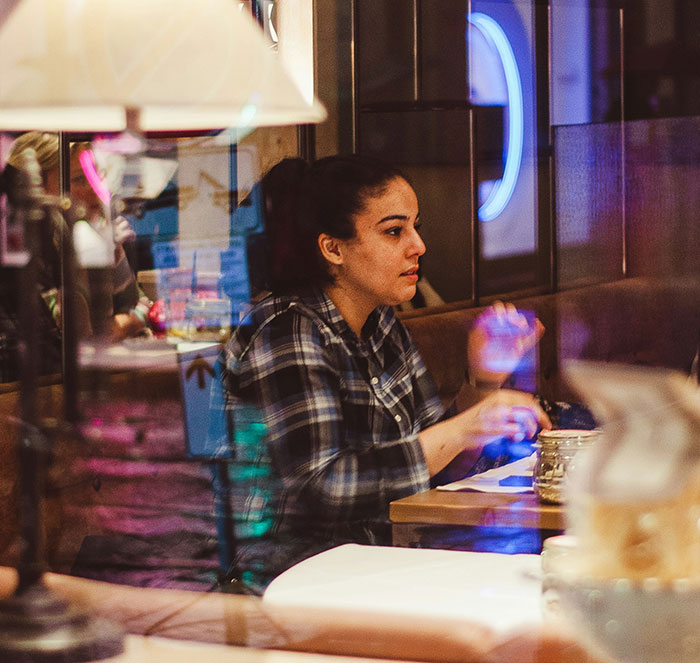 Woman practicing safety habits while seated at a table in a dimly lit indoor setting with reflections on glass.