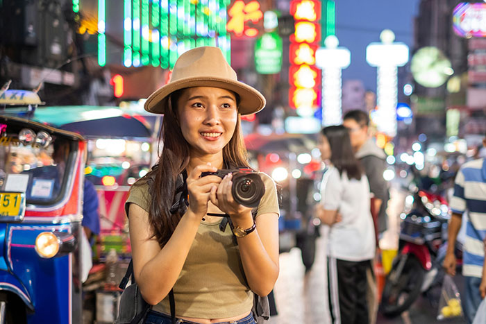 Young woman wearing a hat holding a camera at night in a busy street, illustrating unhinged safety habits concept.