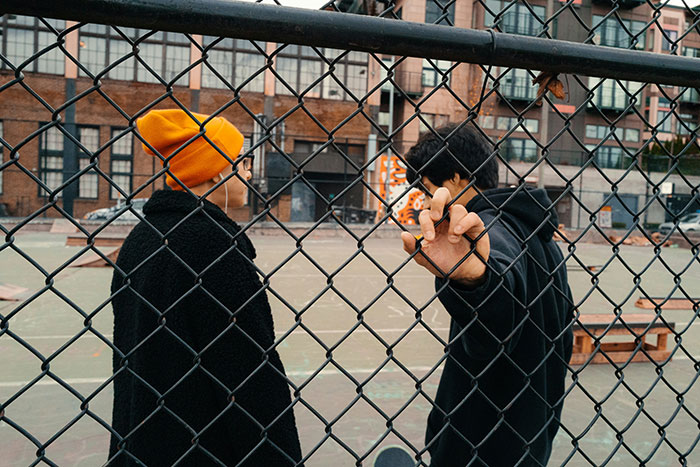 Two women standing by a chain-link fence at an urban basketball court discussing unhinged safety habits.
