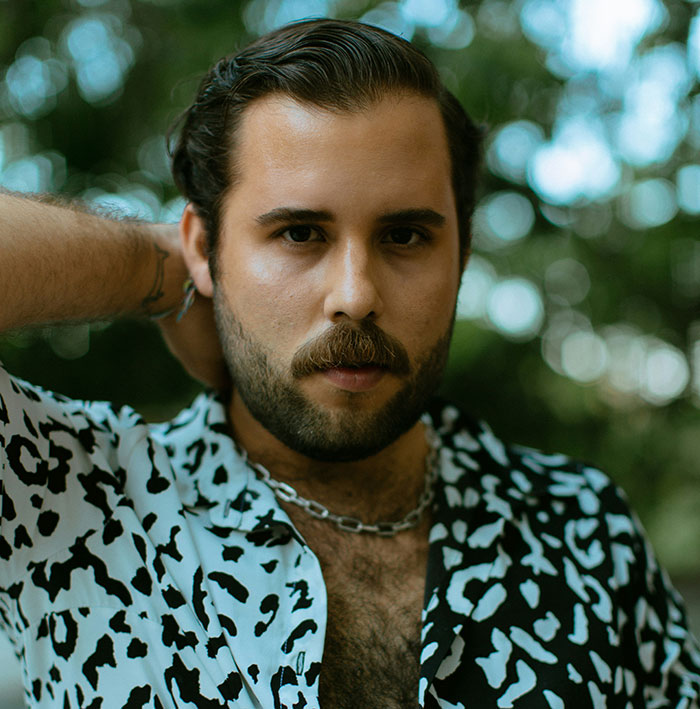 Man with beard and mustache wearing a patterned shirt and chain necklace posing outdoors with a blurred natural background.