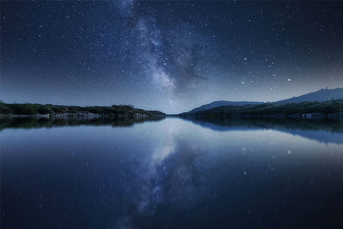 Night sky full of stars reflecting on calm sea waters near a quiet shoreline, capturing eerie maritime calm.