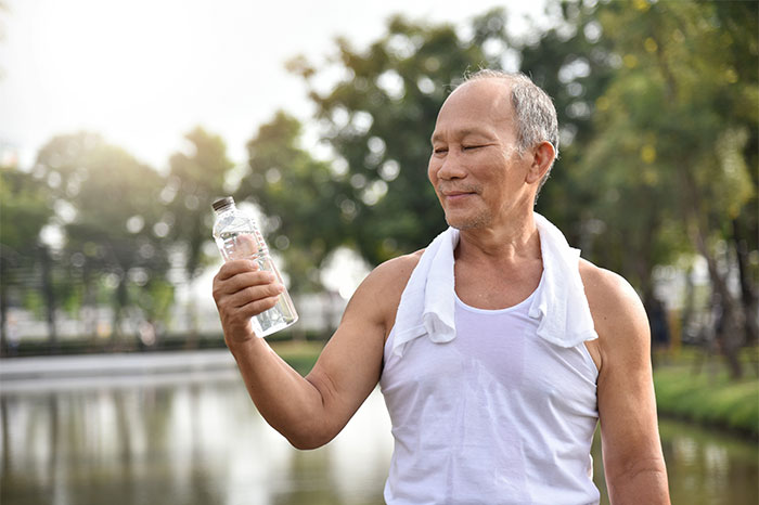Mature man in white tank top with towel around neck holding water bottle outside, showcasing glimpse into world of ultra wealthy.