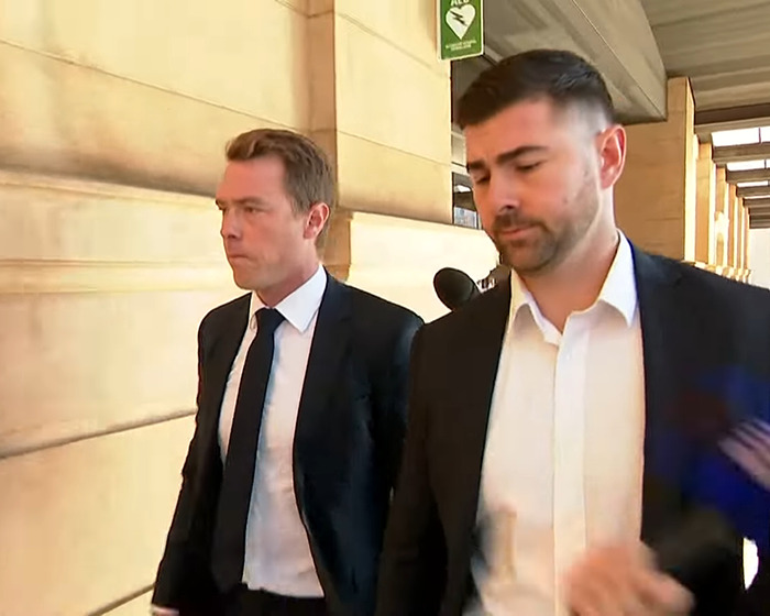 Two men in suits walking solemnly in a courthouse corridor after cyclist pens apology to late Olympian wife.