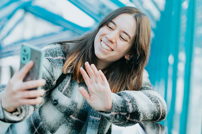 Young woman taking a selfie while smiling and waving, illustrating modern helicopter parenting behavior online.