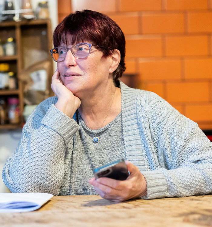 Older woman with glasses sitting at a table holding a phone, depicting a moment of helicopter parenting concern.