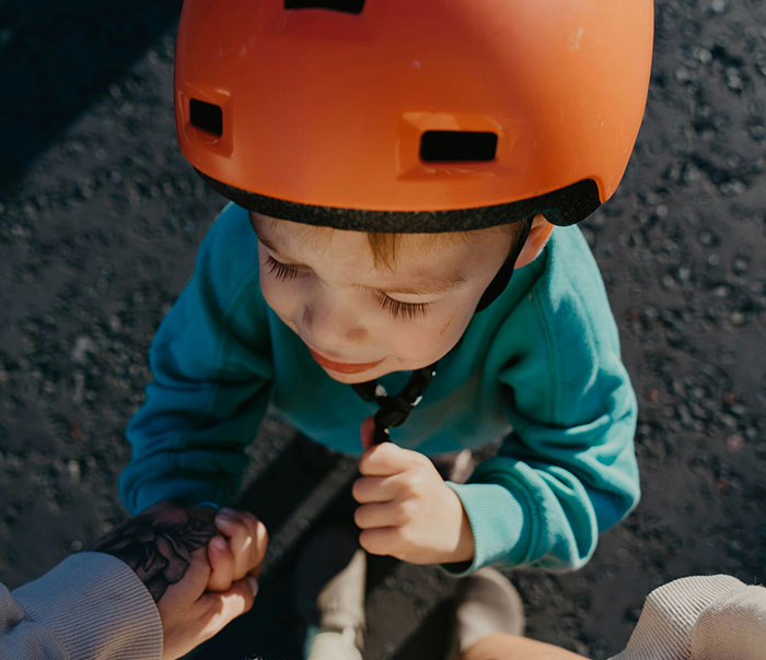Young child wearing an orange helmet holding an adult's hand showing helicopter parenting care and protection outdoors.