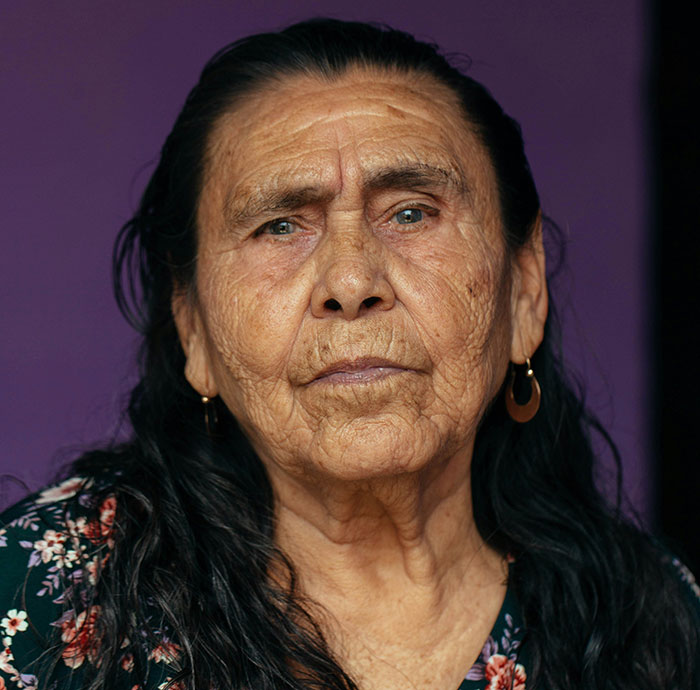 Close-up of an elderly woman with long dark hair and earrings, showing detailed facial wrinkles against a purple background.