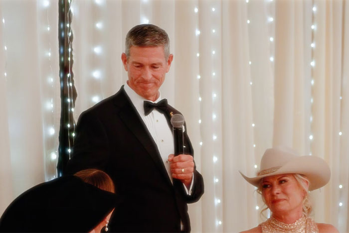 Man in tuxedo holding microphone during chaotic wedding speech, guests dressed in formal and western attire.