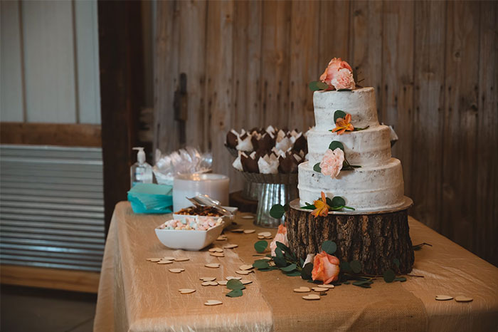 Three-tier wedding cake decorated with flowers on a rustic wooden stand at a wedding chaotic scene table.