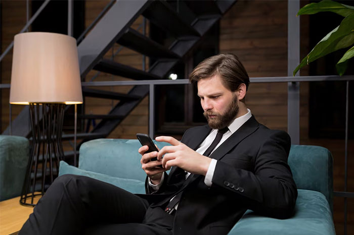 Man in a black suit sitting on a couch, focused on his phone, capturing the chaotic moments at a wedding event.