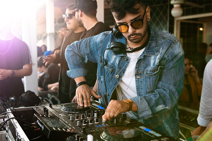 DJ wearing sunglasses and denim jacket mixing music on turntables at a chaotic wedding event with people in background