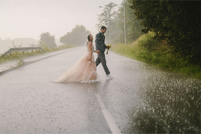 Bride and groom walking on a rainy road, capturing a chaotic wedding moment during an unexpected downpour outdoors.