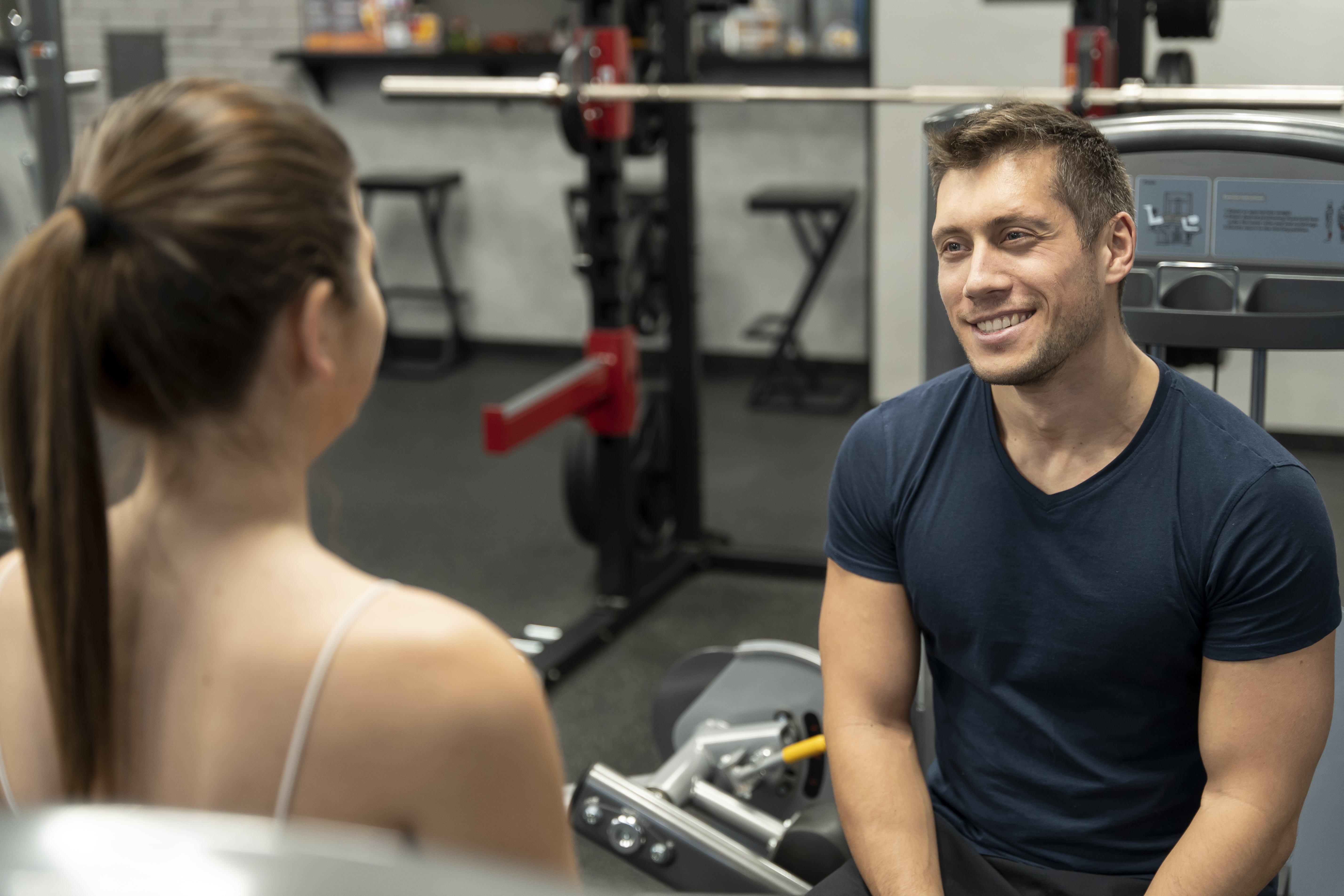 Man watches gym flirting with a woman while smiling, sitting near exercise equipment in a fitness center.