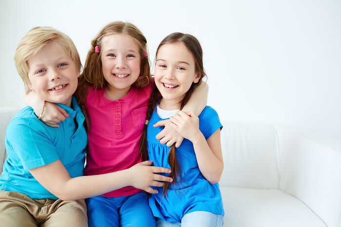 Three happy children sitting closely on a white couch, smiling and showing amusingly bad names reactions.