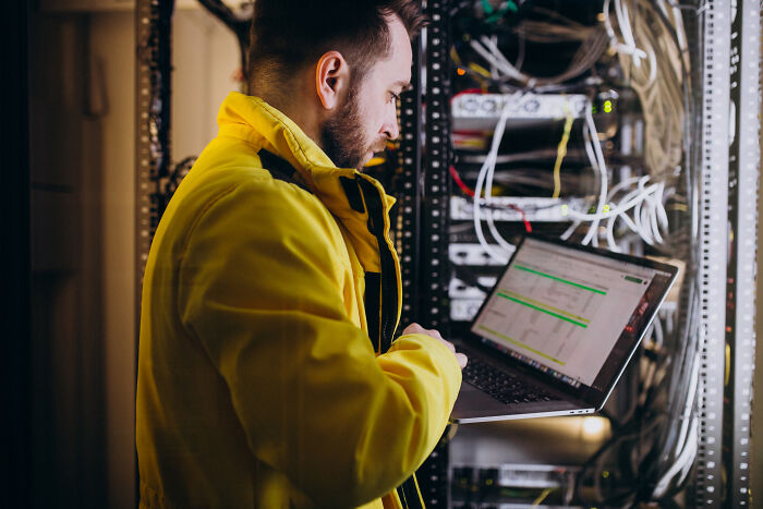 Man in yellow jacket working on laptop in server room managing people share companies bad list data and networks