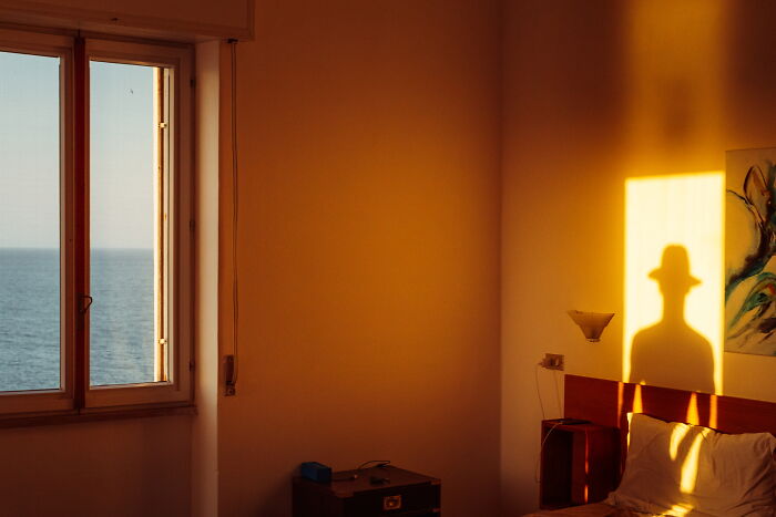 Shadow of a person wearing a hat on a bedroom wall during sunset in a perfectly-timed street photo by Luca Regoli.
