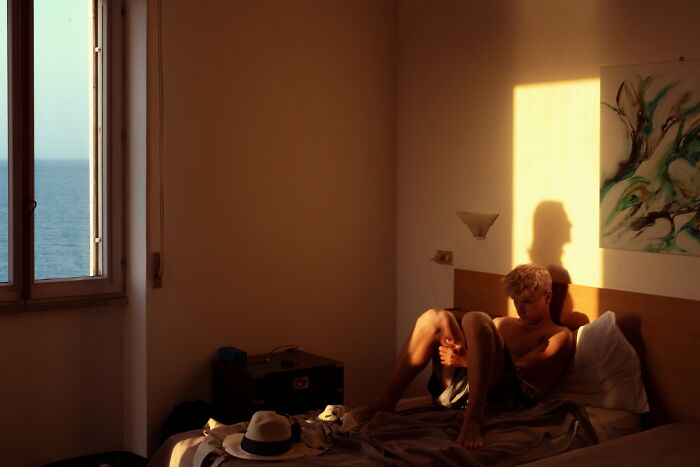 Young man sitting on a bed in warm sunlight, creating a perfectly timed street photo with artistic shadows and calm atmosphere.