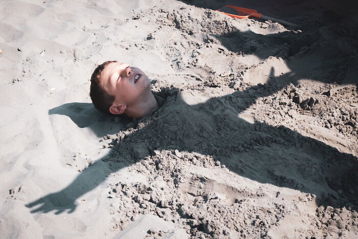Child buried in sand with arms spread wide, captured in a perfectly-timed street photo by Luca Regoli.